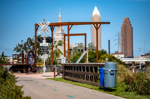 A scenic view of a park with a railroad crossing sign, wooden railing, and city skyline in the background.