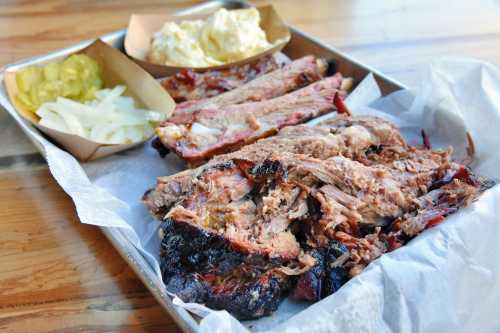 A platter of smoked meats including ribs and brisket, served with pickles, onions, and a side of potato salad.