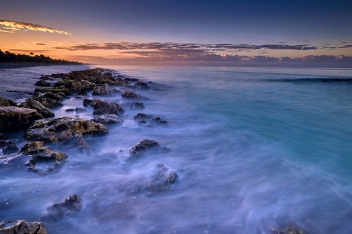 A serene beach scene at sunset, featuring smooth waves gently lapping against rocky shorelines under a colorful sky.