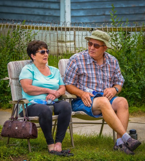 A man and woman sit on lawn chairs, wearing sunglasses, chatting outdoors with greenery in the background.