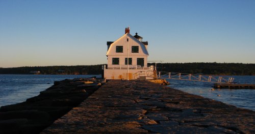 A white lighthouse stands at the end of a stone pier, surrounded by calm water and a clear blue sky.