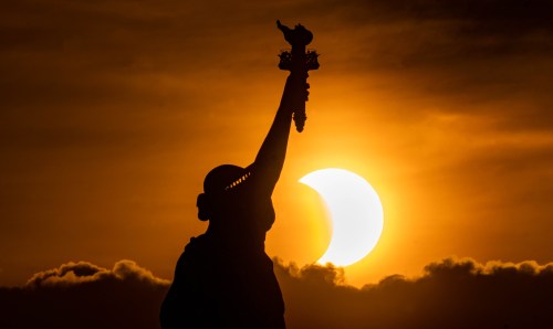 Silhouette of the Statue of Liberty against a sunset with a partial solar eclipse in the background.