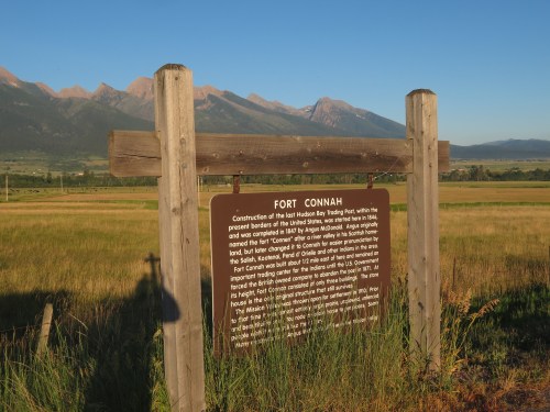 Sign for Fort Connah with mountains in the background, set in a grassy field under a clear blue sky.