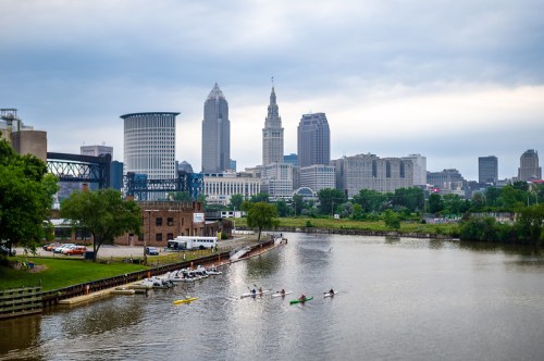 A view of a city skyline with kayakers on a river, surrounded by green trees and overcast skies.