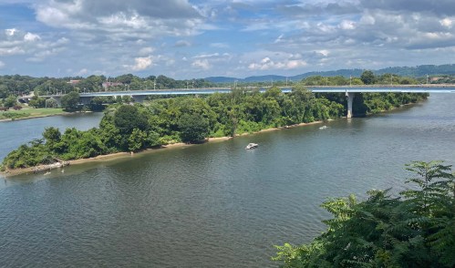 A scenic view of a river with lush greenery, a small island, and a bridge in the background under a partly cloudy sky.
