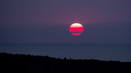 A vibrant sunset over the ocean, with a large sun partially obscured by clouds and a dark silhouette of trees in the foreground.