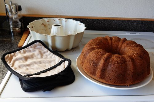 A freshly baked bundt cake on a plate next to a bundt pan and two oven mitts on a kitchen countertop.