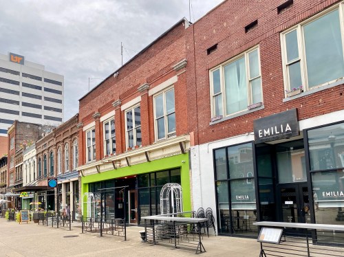 A street view of brick buildings with shops, including "EMILIA," under a cloudy sky in an urban setting.