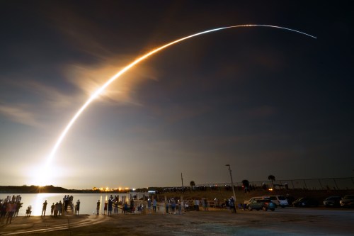 A rocket launches into the night sky, leaving a bright trail, with spectators watching from a nearby shore.