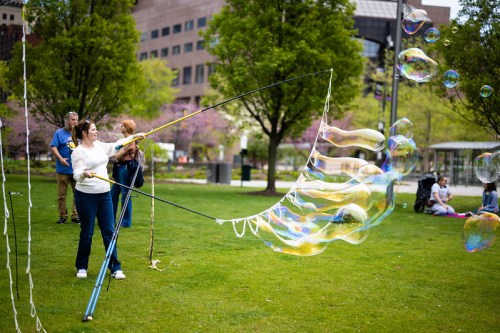 A woman creates large soap bubbles in a park, with people enjoying the scene in the background.