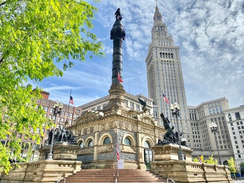 A historic monument with a tall column and statues, surrounded by greenery and modern buildings under a blue sky.