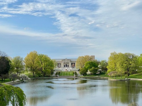 A serene park scene featuring a lake, lush green trees, and a grand building in the background under a blue sky.