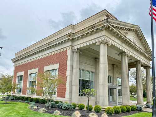 Historic building with columns and large windows, featuring a brick facade and landscaped greenery in front.