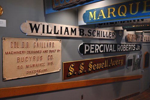 A collection of vintage signs on a wall, featuring names and businesses like "William B. Schiller" and "S.S. Sewell Avery."