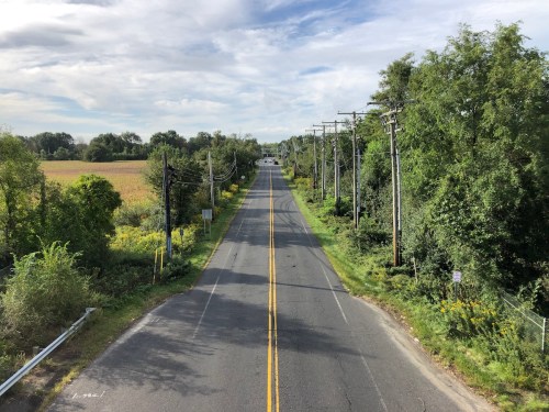 A quiet rural road lined with trees and power lines, leading towards a distant horizon under a partly cloudy sky.