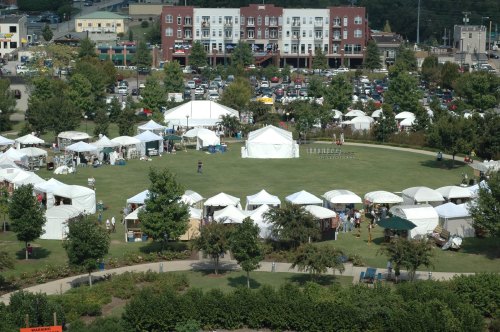 Aerial view of a park with white tents set up for an outdoor event, surrounded by trees and parked cars in the background.