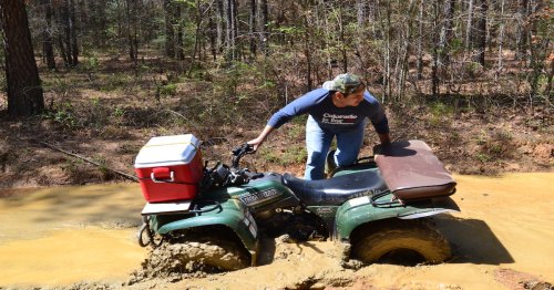 A person struggles to maneuver an ATV stuck in muddy water in a forested area. A cooler is attached to the vehicle.