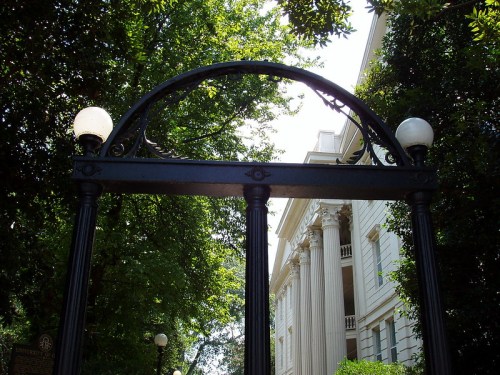 A decorative gate framed by lush green trees, leading to a historic building with columns in the background.