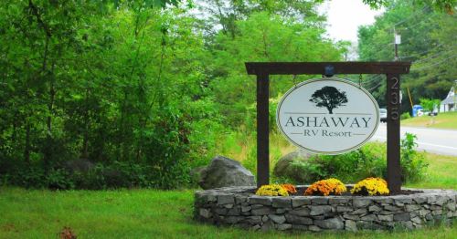 Sign for Ashaway RV Resort surrounded by greenery and flowers, with a road visible in the background.