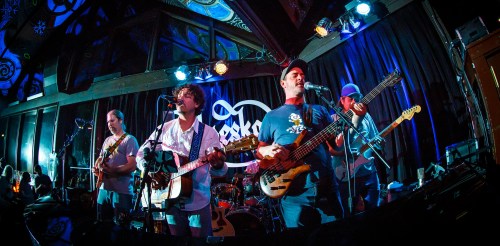 A band performs on stage with instruments, colorful lights, and an engaged audience in a lively venue.