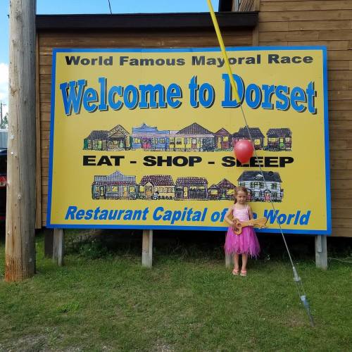A young child stands in front of a colorful sign welcoming visitors to Dorset, promoting eating, shopping, and sleeping.