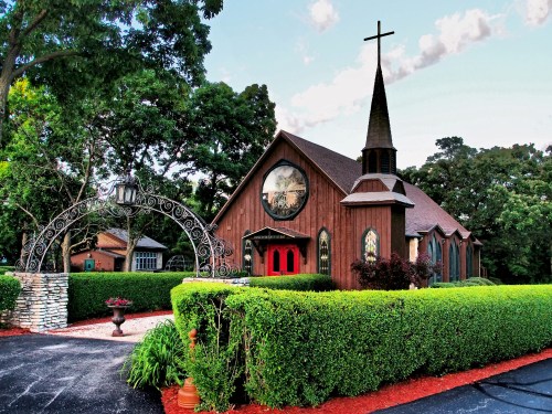 A charming wooden church with a tall steeple, surrounded by lush greenery and colorful landscaping.