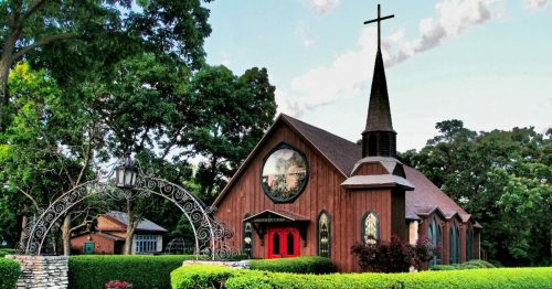 A charming wooden church with a tall steeple, surrounded by lush greenery and colorful landscaping.