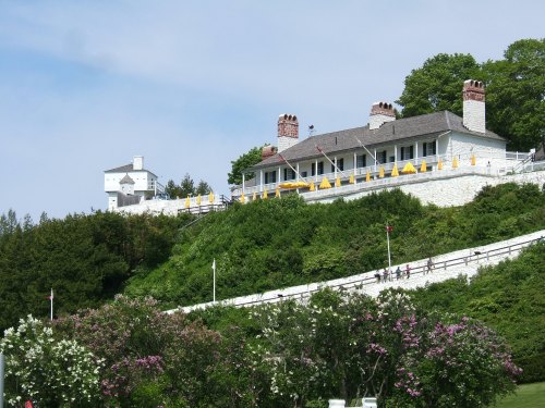 A white building with a porch sits on a hillside, surrounded by greenery and flowers, under a clear blue sky.