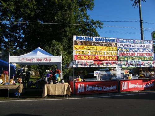 A vibrant outdoor food market with colorful banners for various food stalls, including Polish sausage and funnel cakes.