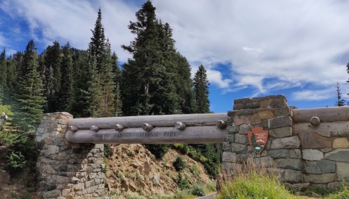 Entrance to a national park featuring a log and stone archway, surrounded by tall evergreen trees and a blue sky.