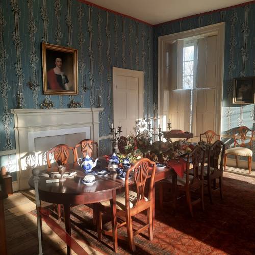 A vintage dining room with a wooden table set, blue wallpaper, and portraits on the walls, illuminated by sunlight.