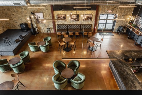 A spacious, modern venue with green chairs, wooden tables, and a stage, featuring stone walls and polished concrete floors.