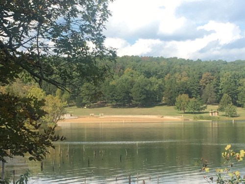 A serene lake surrounded by green trees and a sandy shore under a partly cloudy sky.