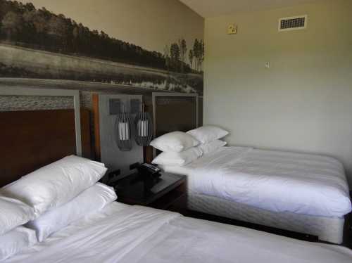 A hotel room featuring two beds with white linens and a scenic wall mural of a forested landscape.