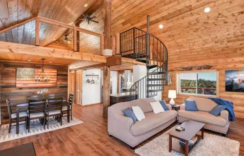 Cozy wooden interior of a cabin with a spiral staircase, living area, dining space, and large windows.