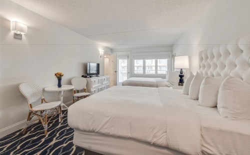 Bright hotel room with two beds, a small table, chairs, and a TV, featuring light decor and a cozy atmosphere.