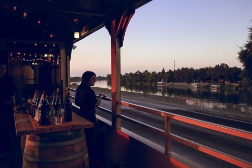 A serene riverside scene at dusk, featuring a person by a bar with bottles, surrounded by trees and calm water.