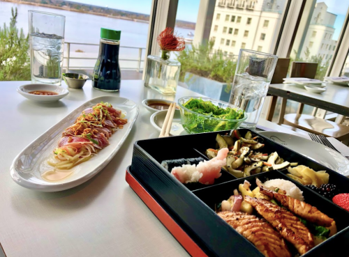 A beautifully arranged sushi bento box with sashimi, salad, and condiments, overlooking a scenic river view.