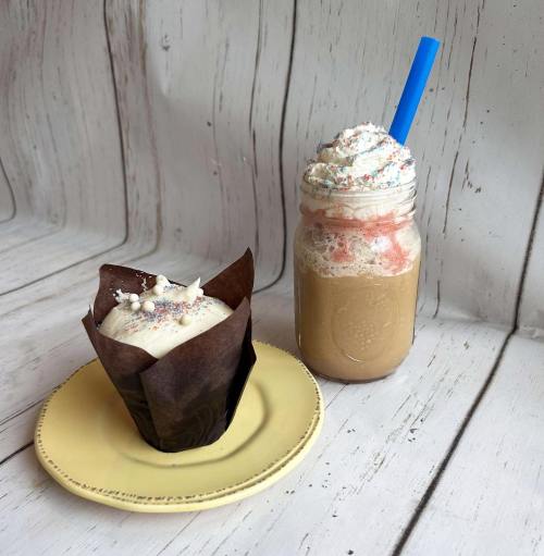 A cupcake on a plate next to a frothy drink in a jar with whipped cream and a blue straw, set against a wooden background.
