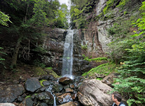 A serene waterfall cascades down rocky cliffs, surrounded by lush greenery and boulders in a tranquil forest setting.