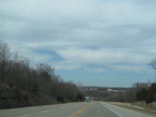A wide, empty road stretches ahead, flanked by bare trees and a cloudy sky in the distance.