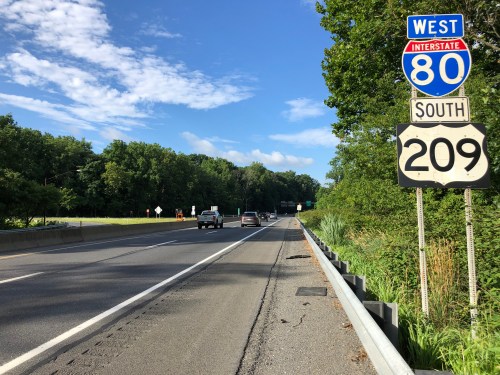 A highway scene showing Interstate 80 West and Route 209 South signs, with cars traveling on a sunny day.