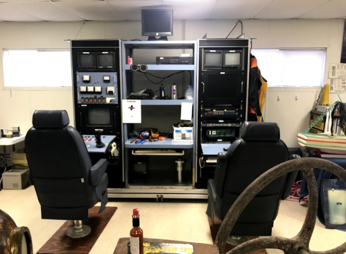 Control room setup with two chairs, monitors, and various equipment on shelves, in a well-lit space.