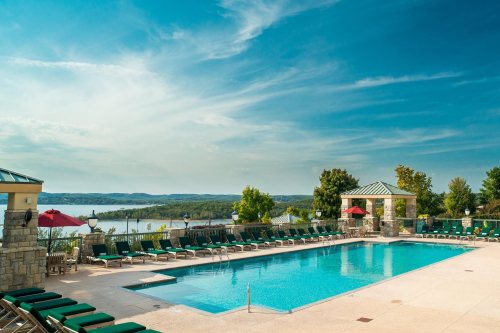 A serene outdoor pool area with lounge chairs, umbrellas, and a scenic view of the lake and trees under a blue sky.