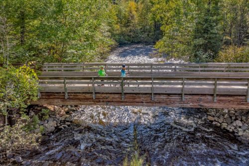 Two people walk on a wooden bridge over a flowing river, surrounded by lush green trees in a scenic landscape.