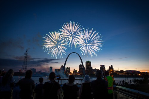 Fireworks burst over the Gateway Arch as spectators watch from the riverfront at dusk. City skyline in the background.