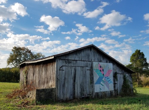 A weathered barn with a colorful quilt pattern on its side, set against a blue sky with fluffy clouds.