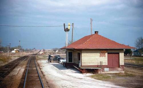 A vintage train station with a red roof, alongside railway tracks and a classic car parked nearby.
