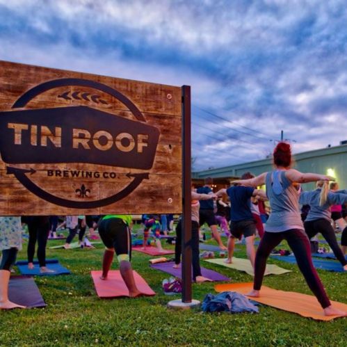 A group of people practicing yoga outdoors in front of a sign for Tin Roof Brewing Co. at dusk.