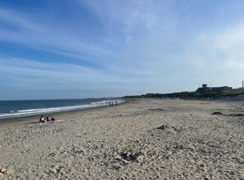 A sandy beach with gentle waves, under a clear blue sky, and a few people walking along the shore.